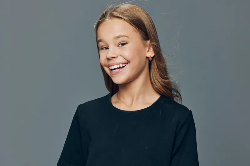Girl smiling and looking at camera with happiness and joy on face against plain background. Young female child with long hair and casual black shirt standing indoors.