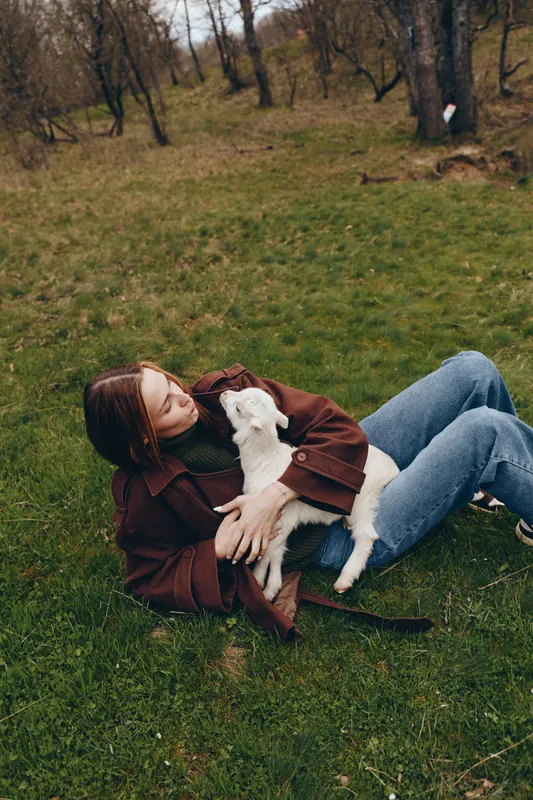 A woman lies on green grass outdoors, cuddling a small white goat kid in a brown coat. A cozy lifestyle moment showing connection with animals in a calm park setting.
