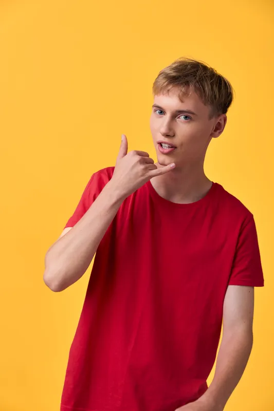 Young man in a red shirt making a call gesture with his hand near his face against a bright yellow backdrop, conveying casual communication and contemporary style