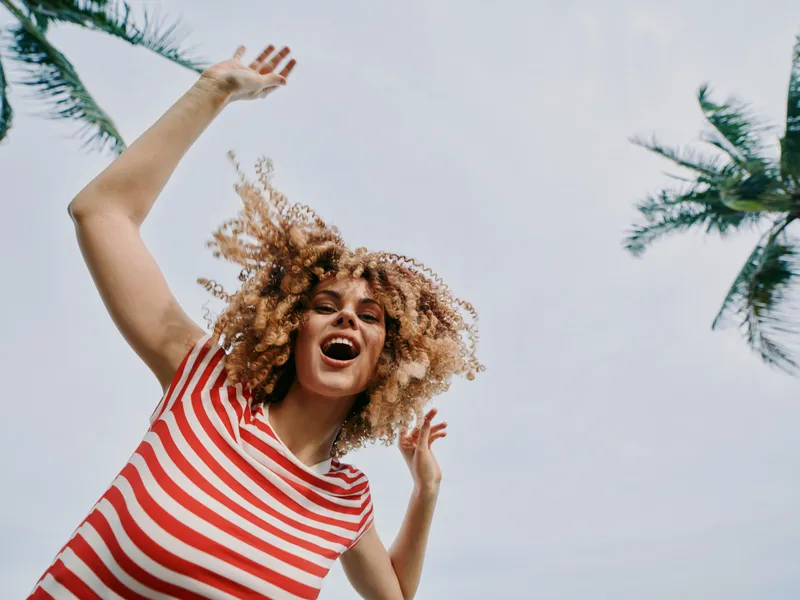 Joyful person jumps outdoors in a red striped shirt, arms raised, smiling broadly against a bright sky with palm trees, capturing carefree energy and playful fashion.