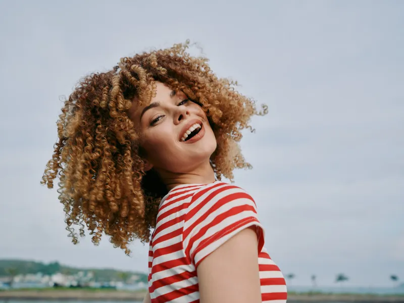 Joyful portrait of a woman with voluminous curly hair wearing a red and white striped shirt, smiling toward the camera outdoors under a bright sky.