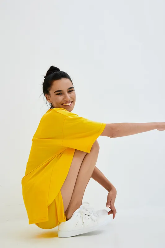 young woman in yellow oversized shirt smiling and squatting on white background, casual style, minimalistic setting, bright colors, youthful energy, studio lighting