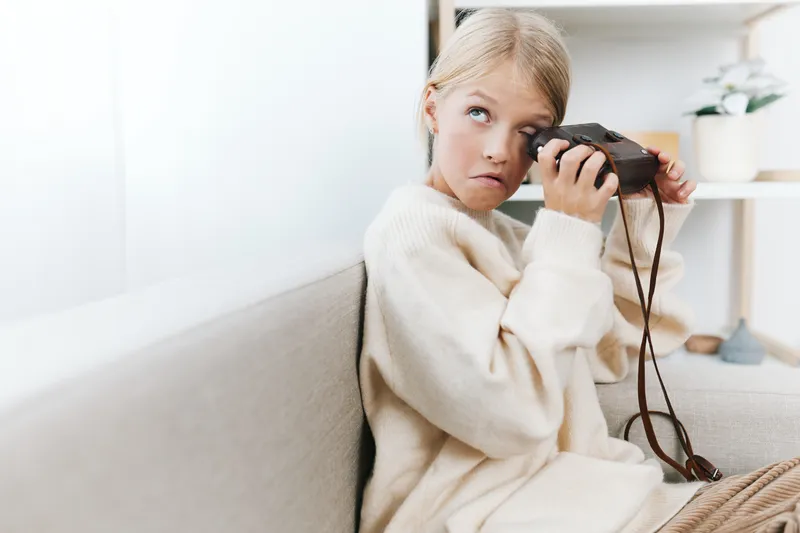 An authentic lifestyle portrait of a young girl indoors, holding a camera with a cozy sweater, capturing a candid moment that highlights everyday school life and playful learning.