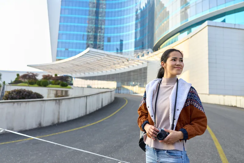 Woman with a camera in hand stands along a modern city street, architectural glass towers above, casual jacket and jeans conveying travel and urban exploration