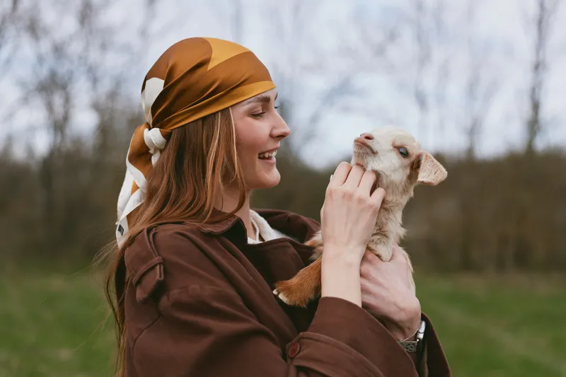A joyful woman in a brown jacket and scarf holds a small goat kid, sharing affection outdoors in a grassy countryside scene, capturing bonding, gentle care, and a warm rural lifestyle.