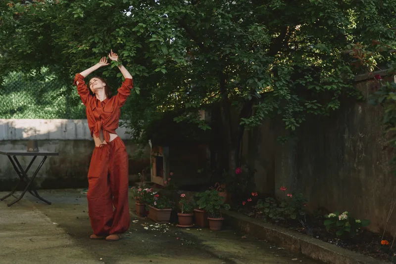 Young woman in a rust red dress dances in a sunlit garden, joyful mood, casual outdoor moment with lush greenery and potted plants behind, bright and lively scene