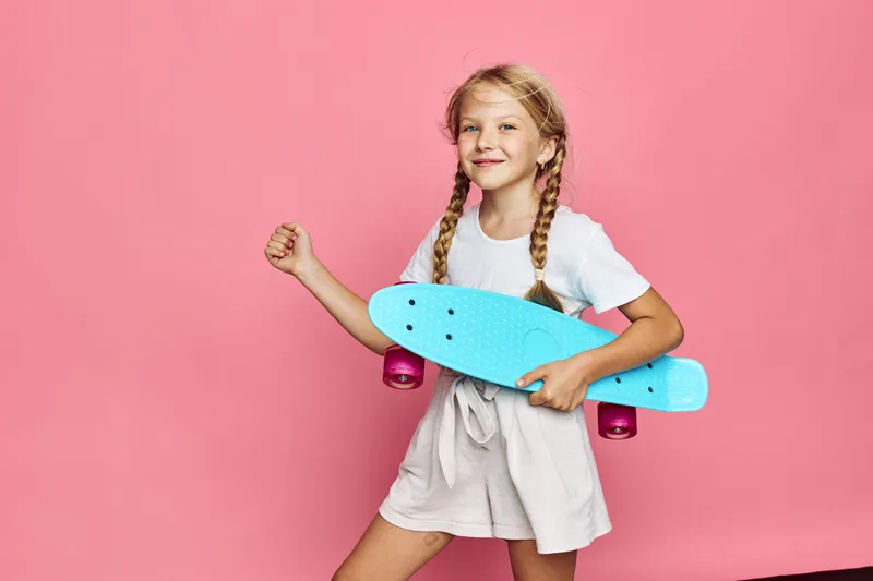 Smiling young girl with braids holds a blue skateboard on a pink background. She wears casual light clothing and poses confidently with a cheerful expression.