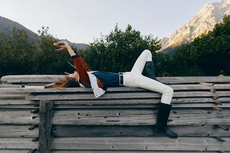 Woman lying on wooden fence relaxing outdoors with mountains in background, casual outfit and boots. Horizontal portrait, peaceful nature scene with trees and golden hour glow.