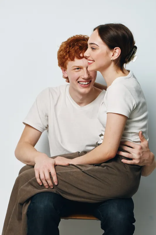 Young couple sitting together smiling and embracing with happiness casual clothing and relaxed pose against white background in studio setting.