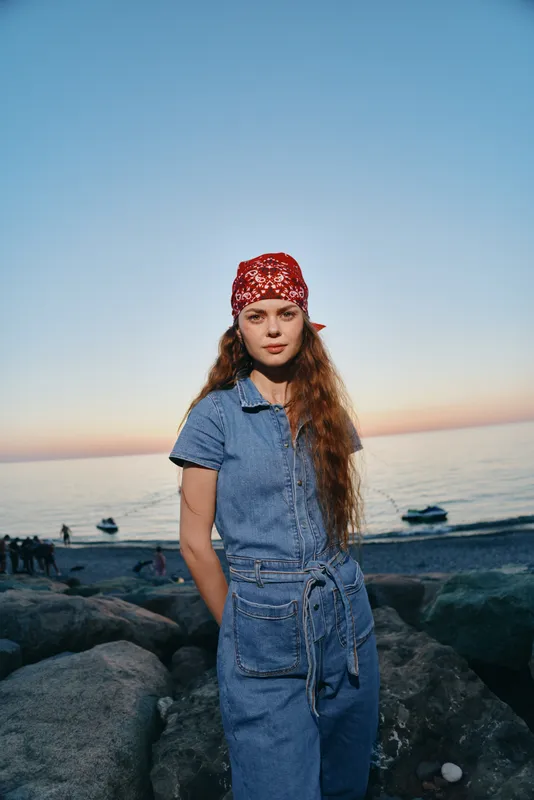 young woman wearing denim jumpsuit and red bandana standing on rocky shore near calm sea at sunset with soft colorful sky