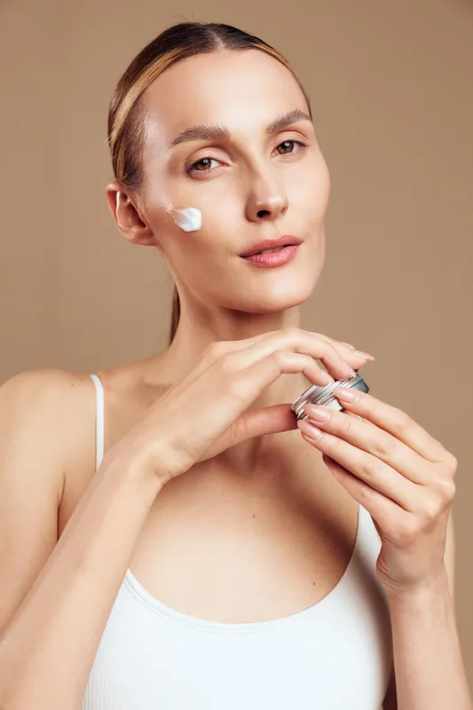 A woman in a white tank top applies cream on her face with a calm expression, holding a jar of skincare product. The beige background emphasizes natural beauty and self-care routine.