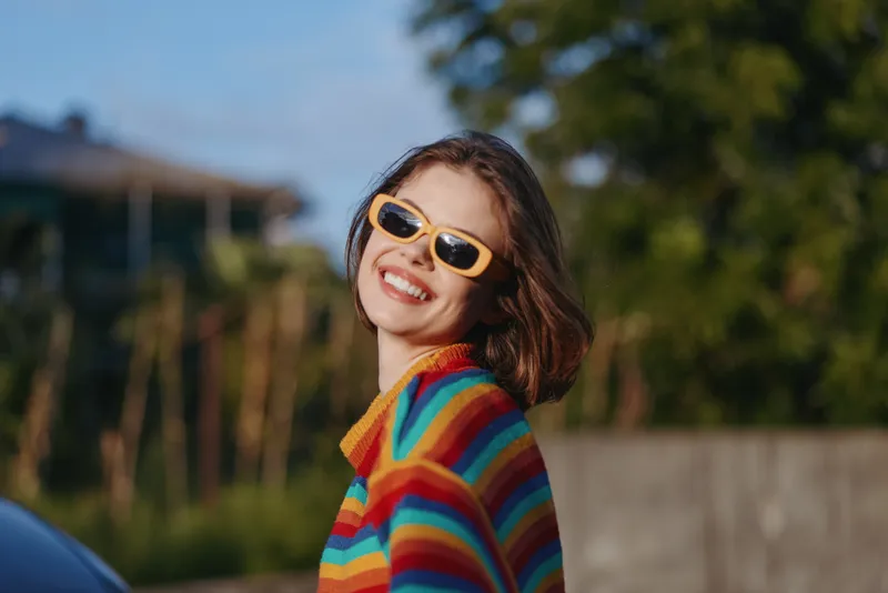 woman smiling striped sweater yellow sunglasses young adult outdoor lifestyle fashion, early twenties model turning head in sunlit portrait, colorful casual outfit for travel and street style