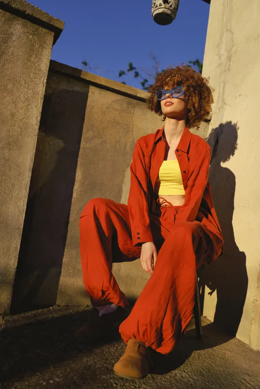 Confident woman in bold red suit and yellow top poses outdoors against a concrete wall, showcasing contemporary fashion lifestyle and sunny, playful mood