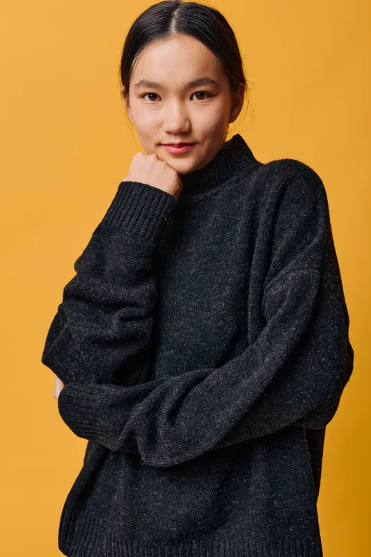 Young woman standing with hand near chin wearing dark sweater on orange background, confident and thoughtful expression portrait in studio with simple colorful backdrop.