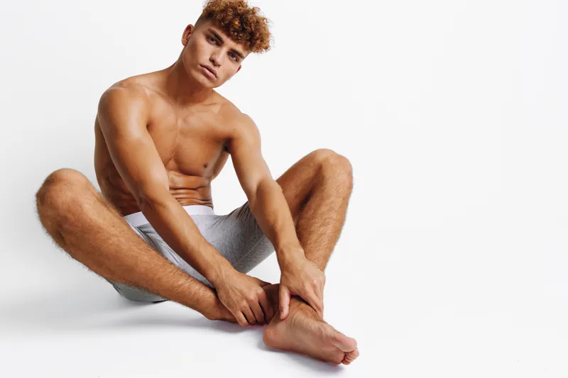 Man with muscular body sitting on white floor with legs stretched wearing gray shorts and looking at camera in studio, relaxed and confident pose with curly hair.