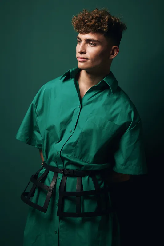 Young man with curly hair wearing oversized police inspired outfit in green attentively looking to side over dark green studio background, posing in fashion natural light portrait.