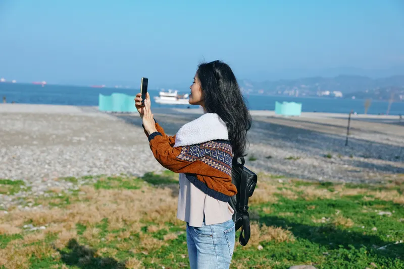 Woman taking photo on a sunny beach with phone held up, windswept hair, casual jacket, jeans, coastal landscape, distant boats, calm sea, vibrant sky, candid travel moment.
