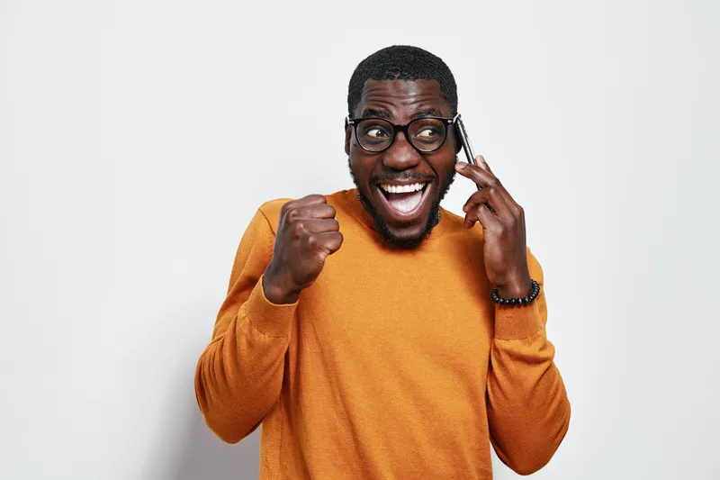 African American man with dark skin wearing glasses and orange sweater talking on phone with excited expression and fist pump gesture on white background.