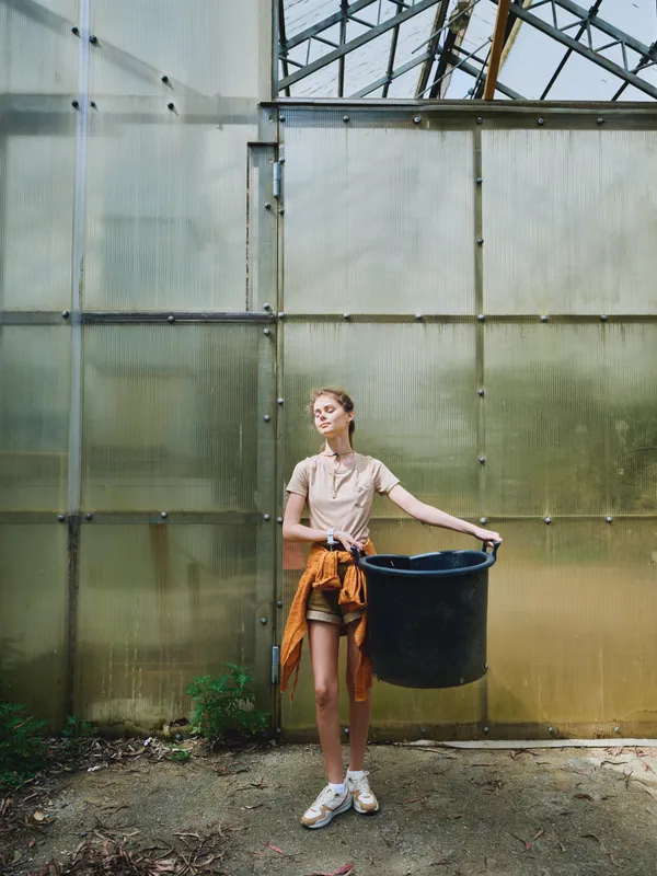 Woman greenhouse gardening with a black bucket container and potted plants, casual outfit with orange jacket tied, standing in glass structure with sunlight, urban plant care scene.