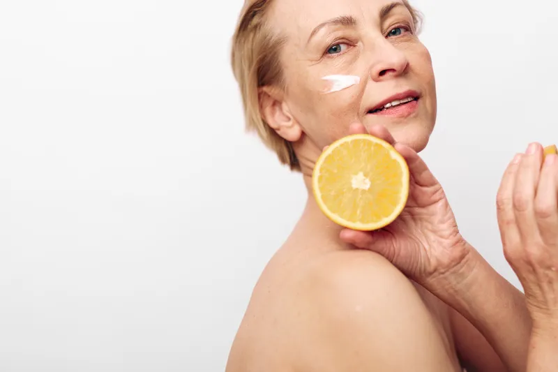 Mature woman with natural skin holds fresh orange slice near face with cream on cheek, smiling gently on white background. Concept for skincare, natural beauty, and healthy lifestyle.