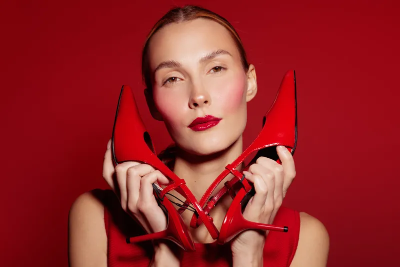 Close-up of a woman holding red high heels near her face against a red background. Concept of fashion, style, elegance, and beauty with vibrant color contrast and confident expression.