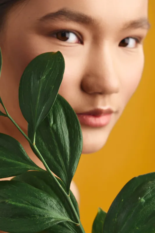 Asian woman appears in a close portrait with vibrant green leaves, warm soft lighting, calm and serene mood, highlighting natural beauty and artistic botanical composition.
