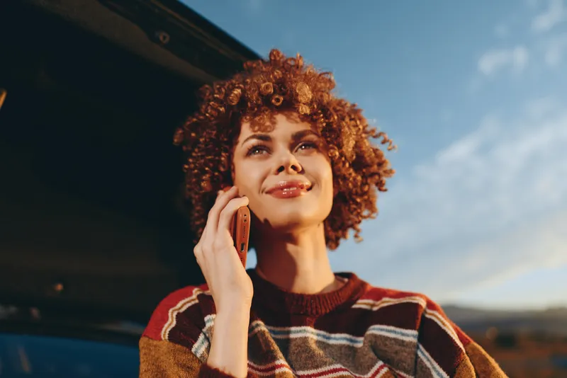 Woman lifestyle portrait outdoors, curly hair, smiling as she holds smartphone for calling on phone, vehicle interior softly blurred, warm daylight sky behind, casual sweater, relaxed