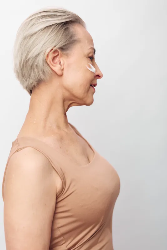 Profile of a mature woman with short blonde hair applying white cream on her cheek. She wears a beige tank top against a plain light background. Concept of skincare, aging, and self-care.