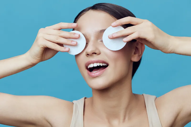 A joyful young woman applying skincare with cotton pads against a blue background, embodying beauty, self care, and freshness in a modern lifestyle