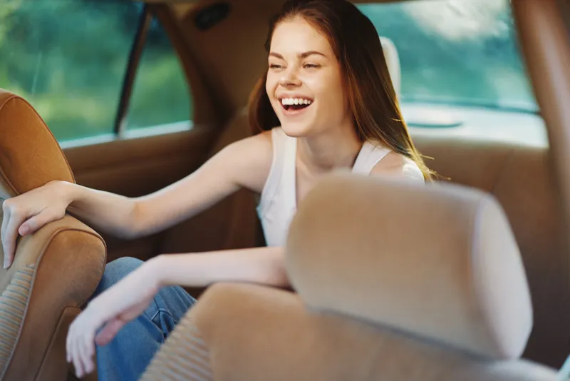 Happy young woman laughing in a car, wearing a casual outfit, showcasing joy and spontaneity amid a bright, vibrant background.