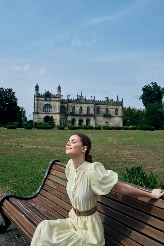 A woman in a light dress sits on a wooden bench in a green park, with a grand castle in the background, enjoying a peaceful serene outdoor moment