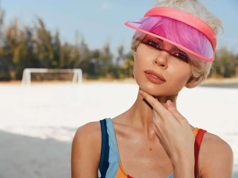 Stylish sunlit portrait of a woman wearing a vibrant pink visor on a sandy beach, posed with a thoughtful smile and a gentle hand touch, embodying summer fashion.