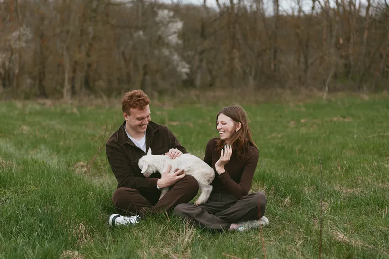 A cheerful couple sits on a grassy field, smiling at a tiny white goat kid resting in their lap. A lifestyle moment capturing joy, companionship, and animal friendship.
