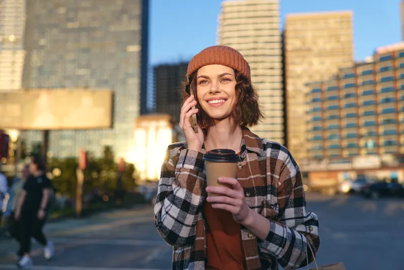 woman holding coffee and phone in urban street, smile in golden hour glow, candid lifestyle portrait emphasizing authenticity, mindful living and emotional storytelling in city scene
