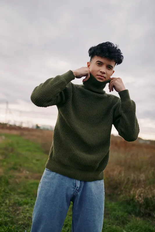 A candid portrait of a young man wearing a green knit turtleneck, standing in an open field. Natural light enhances authenticity and credibility in the shot.
