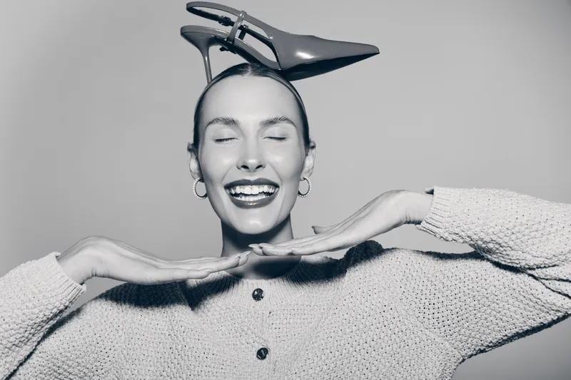 Happy Caucasian woman with fair skin smiling eyes closed holds hands under chin and high heel shoe on head.