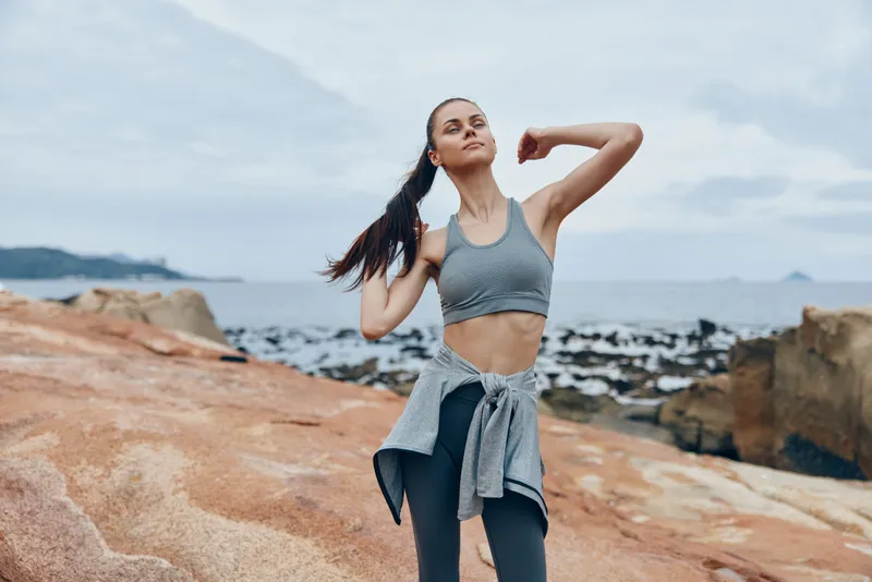 Fit young woman stretching on rocky beach in athletic wear, showcasing healthy lifestyle and fitness, ocean background, cloudy weather, and serene atmosphere