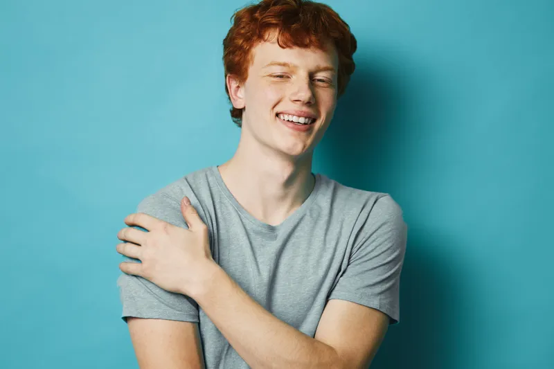Young man with red hair and casual gray t shirt smiling with eyes closed against blue background. Portrait of happy male with relaxed pose and natural expression.