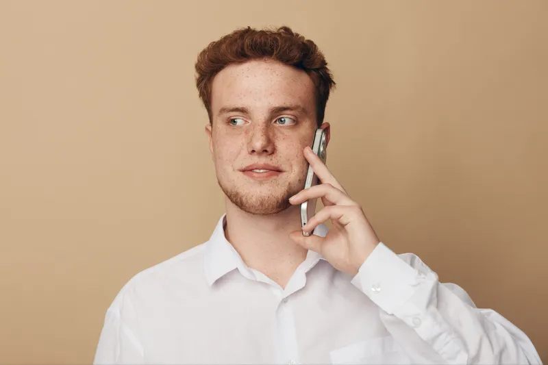 Young man with red hair and freckles wearing white shirt talks on smartphone looking sideways on beige background. Concept of communication, technology, and casual lifestyle.