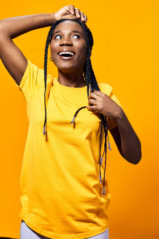 Joyful young Black woman with braided hair in a bright yellow shirt, expressing happiness against a vibrant yellow background, perfect for wellness and positivity themes
