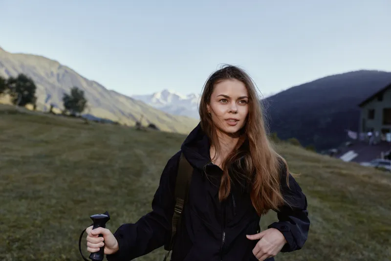 A cheerful woman hiker stands in a scenic outdoor landscape, smiling softly while gripping a trekking pole. She appears confident and prepared, radiating energy, resilience, and adventurous spirit.