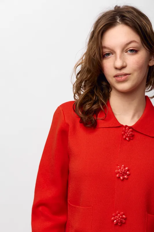 Young woman with curly brown hair wearing a red sweater with decorative buttons poses against a white background. Casual style and natural expression concept.