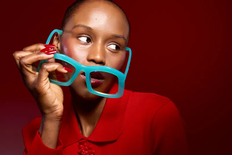Stylish black woman with dark skin holds blue glasses near face, wearing red sweater. Red background highlights fashion and beauty concept with confident expression and colorful accessories.