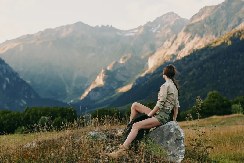 One woman sits on a rock in a mountain meadow, gazing toward distant peaks, conveying calm, freedom and solitude in a pristine outdoor scene.