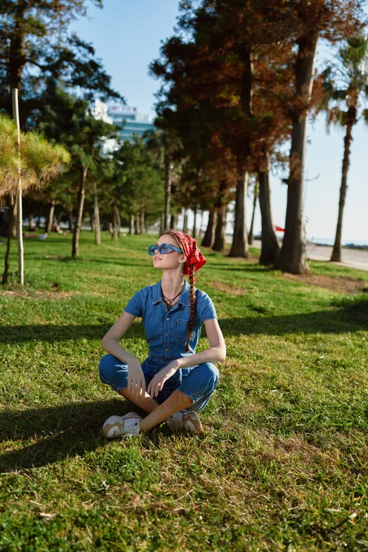 lifestyle woman in modern boho-western denim street style sitting cross-legged on grass under tall trees with sunlight and film color effect, wearing sunglasses and red bandana