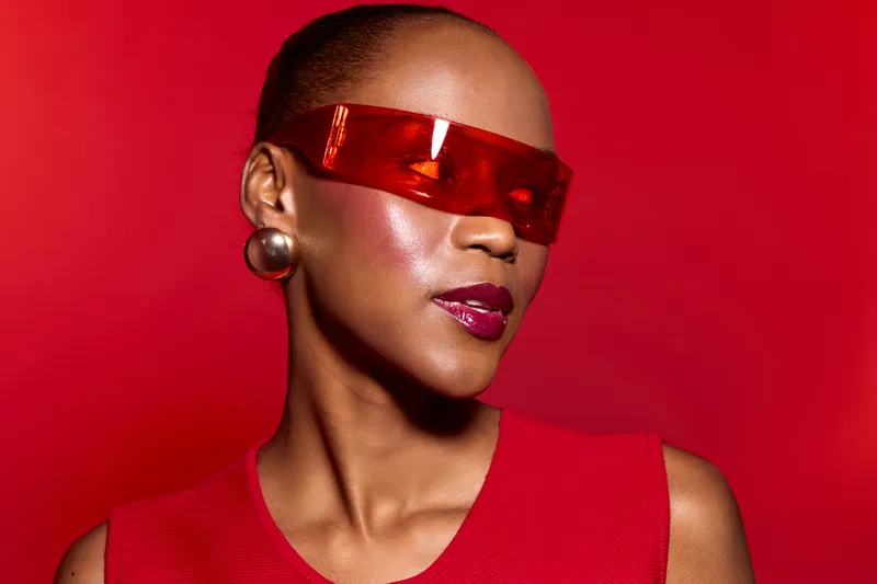 Stylish black woman with dark skin wears red futuristic sunglasses and matching red top against vibrant red background. Concept of fashion, confidence, and modern beauty.