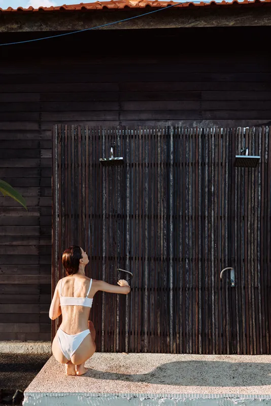 Beach scene with a woman in a white swimsuit kneeling by a rustic wooden door, water spraying from a shower, outdoor lifestyle and relaxation under warm sun.