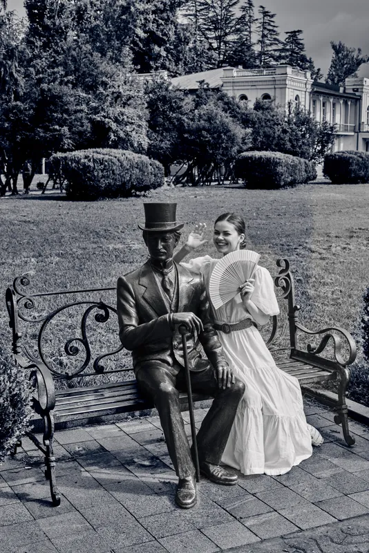 Vintage couple sits beside a statue on an iron bench in a formal garden under trees and arches, the woman in flowing dress with a fan, creating a timeless portrait