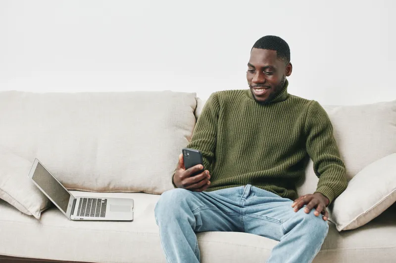Happy young man in green sweater checking smartphone on the couch, representing technology and leisure activities at home in a cozy environment