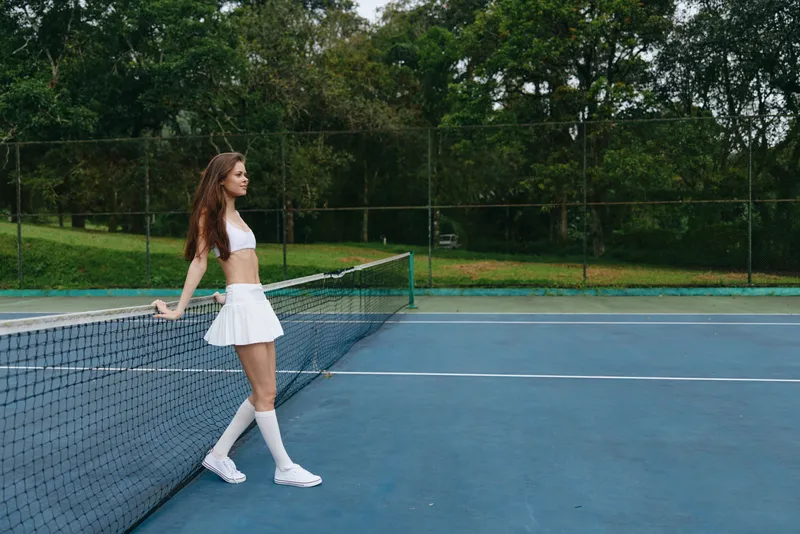 Young woman in a white tennis outfit looking confident while resting on the tennis court, surrounded by lush greenery and a vibrant blue background